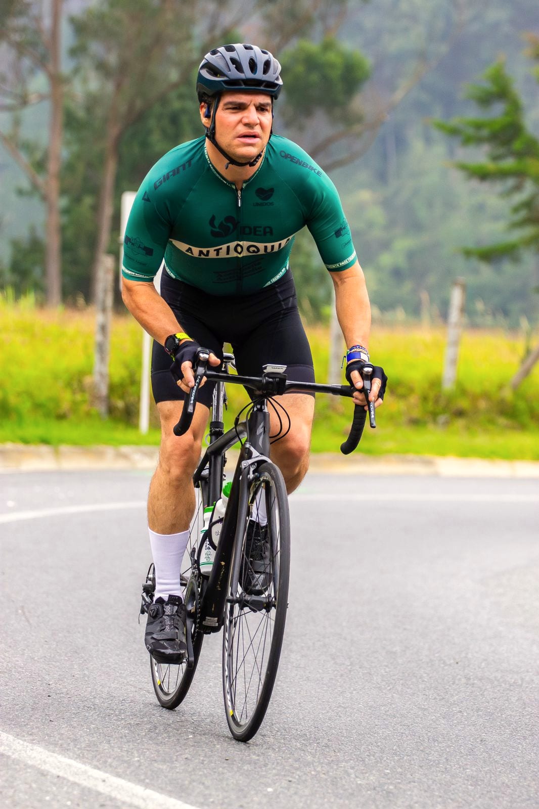 Alejandro Arbeláez A. sube una montaña en bicicleta de ruta, con camiseta verde y casco, en una carretera entre pastizales